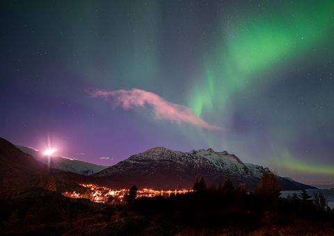Vibrant green aurora borealis illuminates a snow-covered mountain valley with glowing village lights below, creating a magical Arctic night scene.