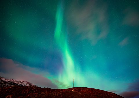 Vibrant northern lights dance across the Arctic sky above a snow-covered mountain ridge with an illuminated radio tower.