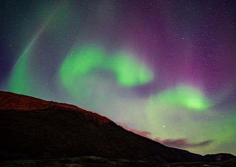 Vibrant green and purple aurora borealis dancing across the night sky above a dark mountainous landscape, with stars twinkling overhead.