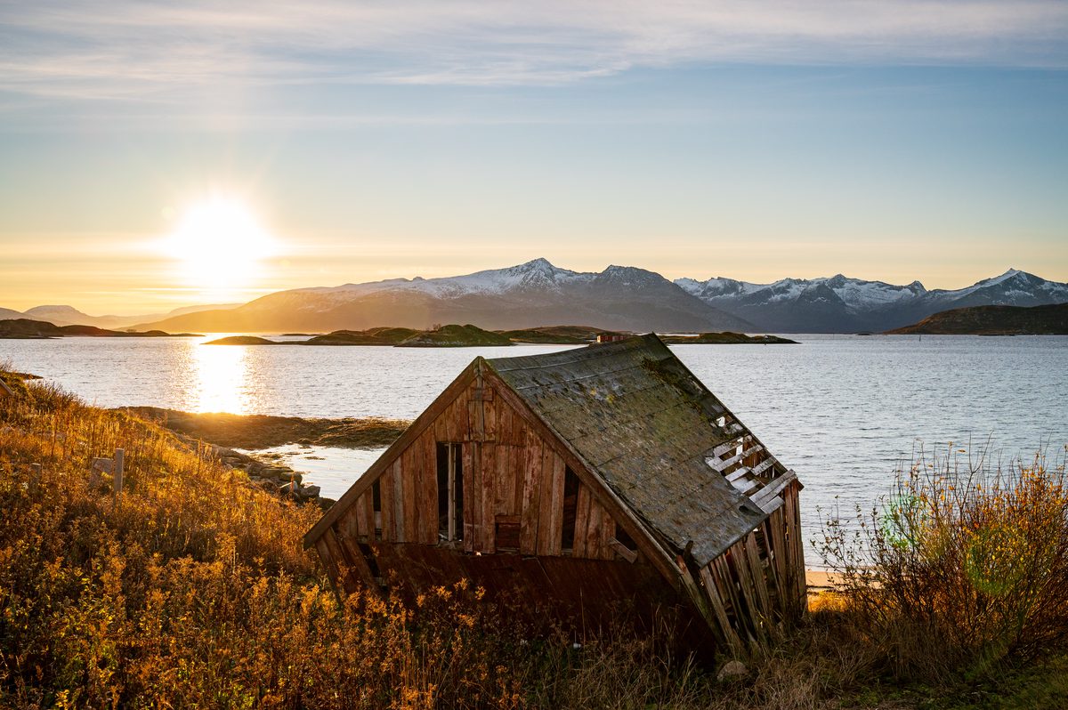 A weathered wooden boathouse stands among autumn vegetation beside a calm fjord in Northern Norway. The low Arctic sun illuminates the water and snow-covered mountains with warm golden light.