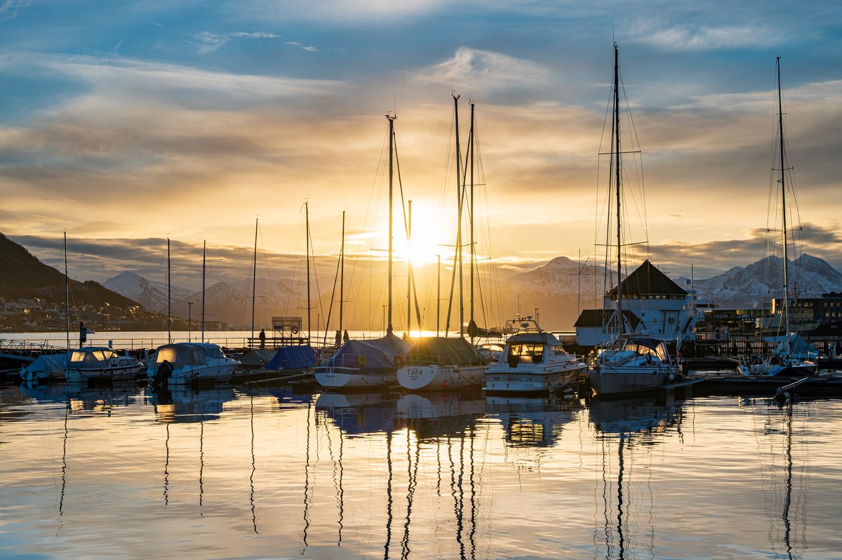 Sailboats and yachts rest on perfectly calm water as the low evening sun sets behind the mountains surrounding Tromsø. Golden reflections and silhouetted masts create a peaceful maritime scene during the Arctic autumn.