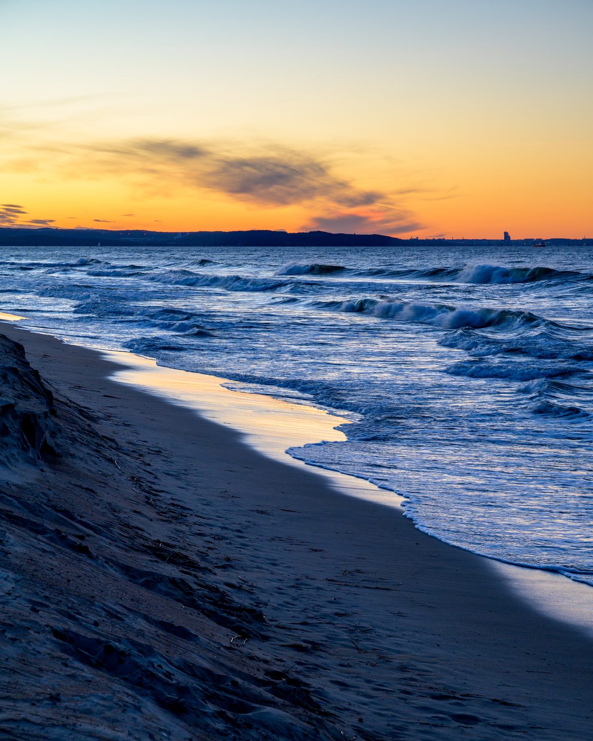 Rolling waves approach the darkened beach beneath a vivid orange and blue sunset. The last light of day catches the wet shoreline, creating a glowing line between land and sea.