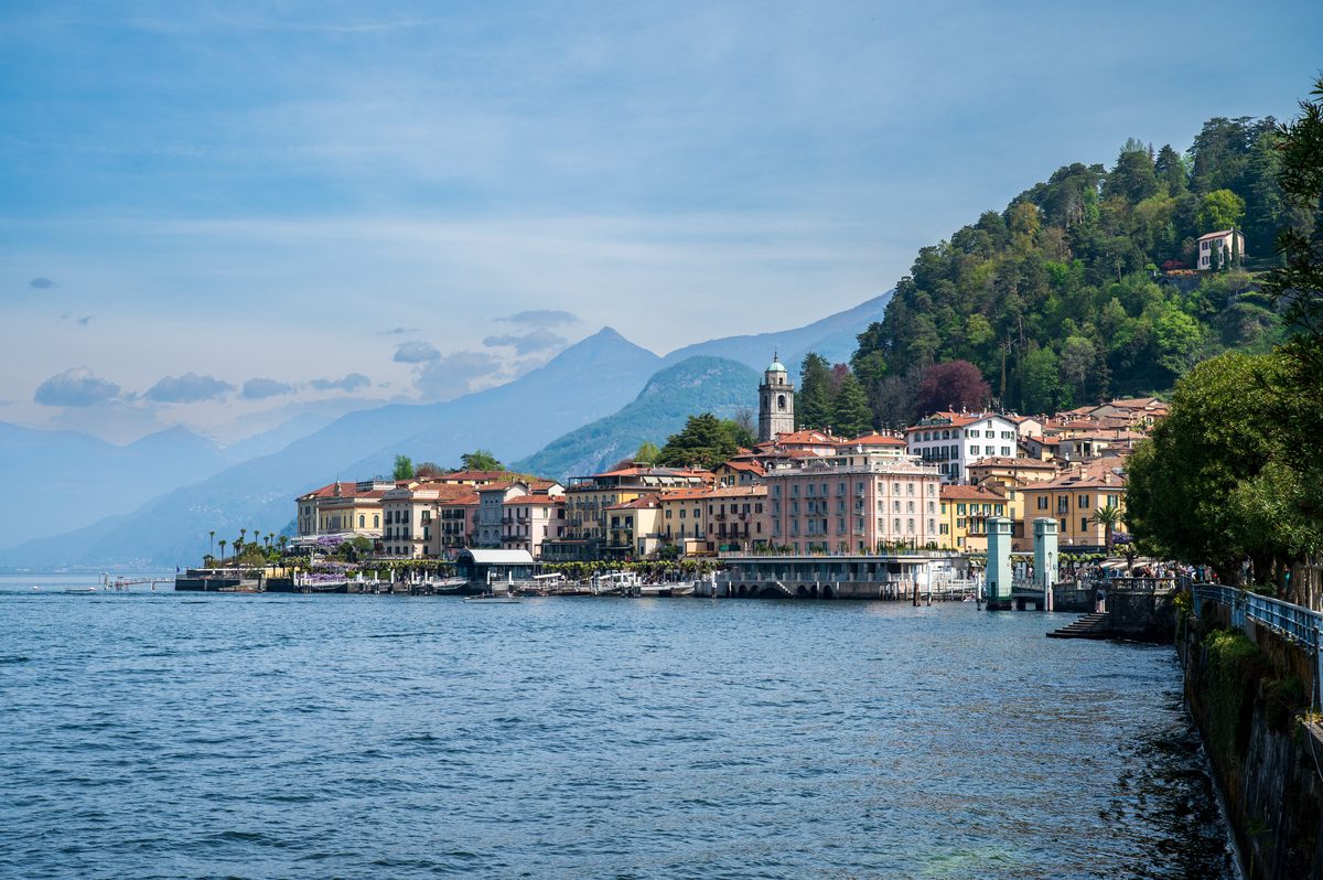 Colourful historic buildings line the waterfront of Bellagio, surrounded by lush hills and distant mountain peaks. The elegant lakeside architecture and tranquil blue water create one of the most iconic views of Lake Como.