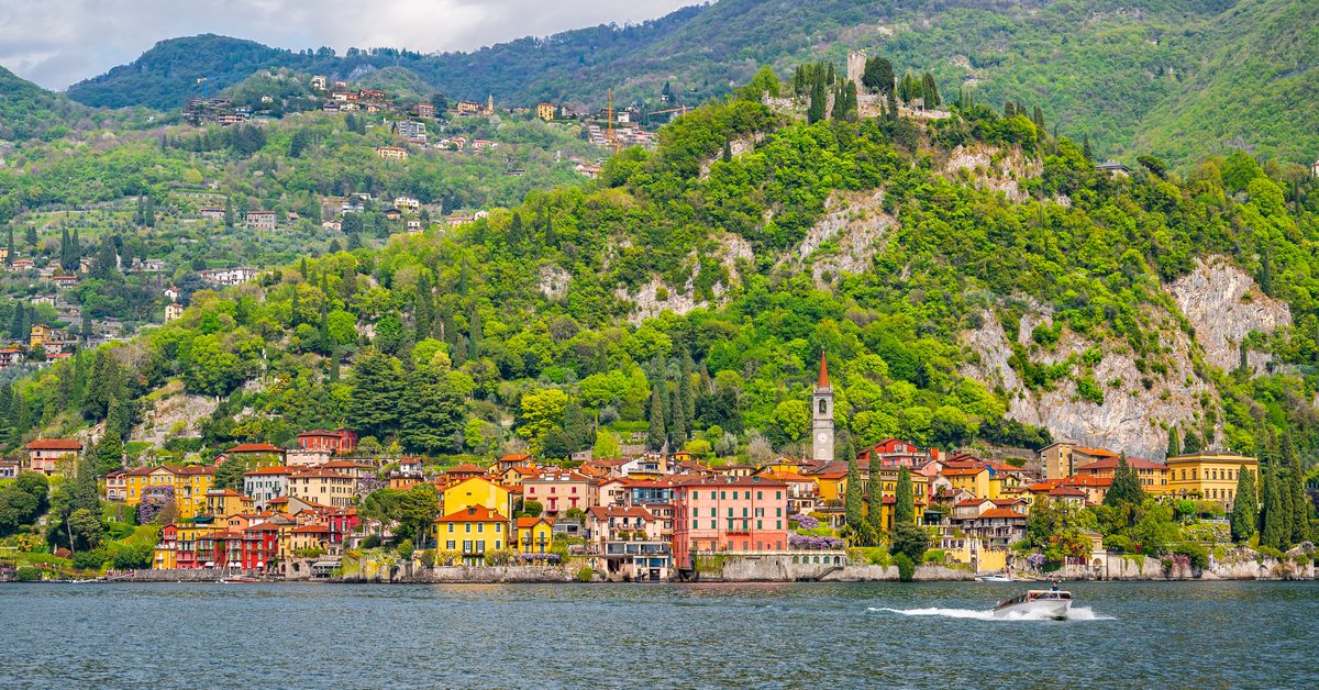 The colourful lakeside village of Varenna stretches along the shore beneath densely wooded mountains and the historic ruins above. A speedboat crossing the foreground adds movement to the otherwise peaceful landscape of Lake Como.