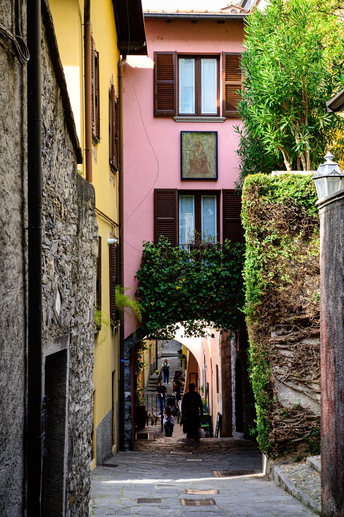 1. A narrow stone passage winds between colourful houses, wooden shutters and cascading greenery in a historic Lake Como village. The intimate architecture and shaded pathway capture the quiet charm of everyday life away from the busy waterfront.