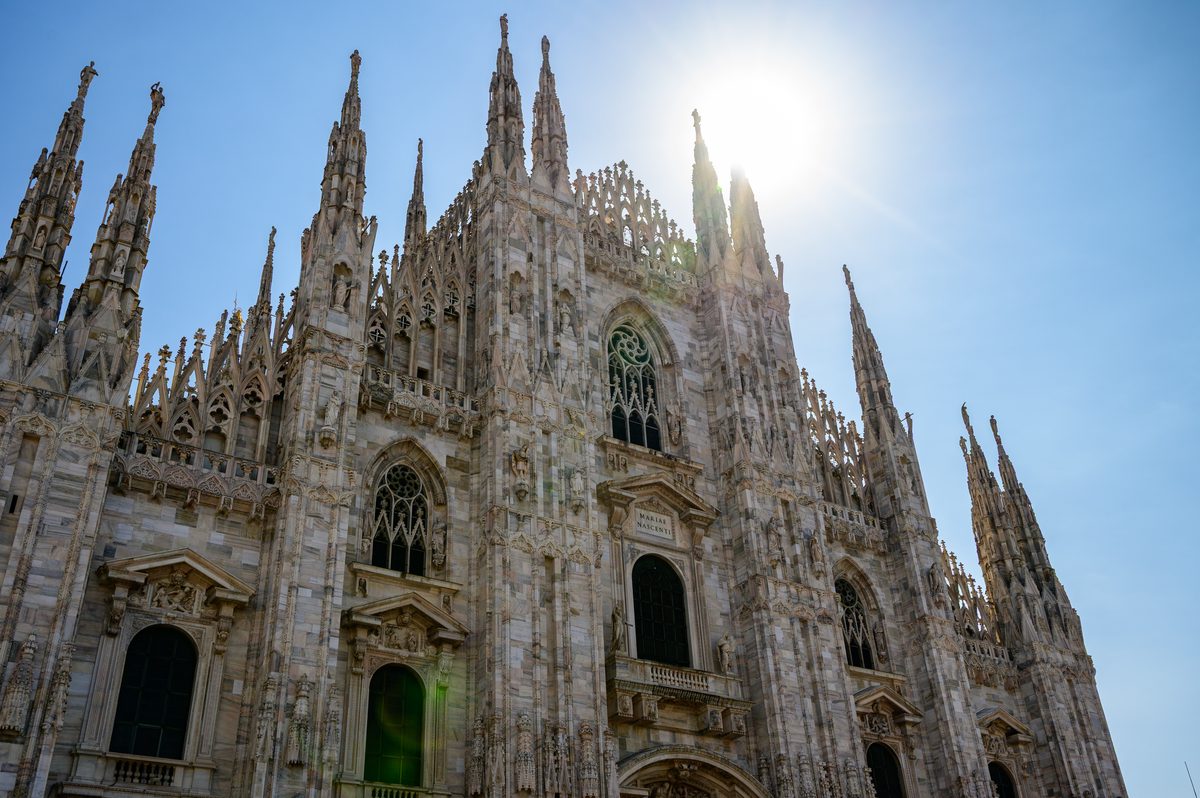 The spring sun shines above the soaring Gothic spires of the Duomo di Milano, illuminating the cathedral’s intricate marble architecture. The low-angle perspective emphasizes the extraordinary scale and detail of Milan’s most famous landmark.