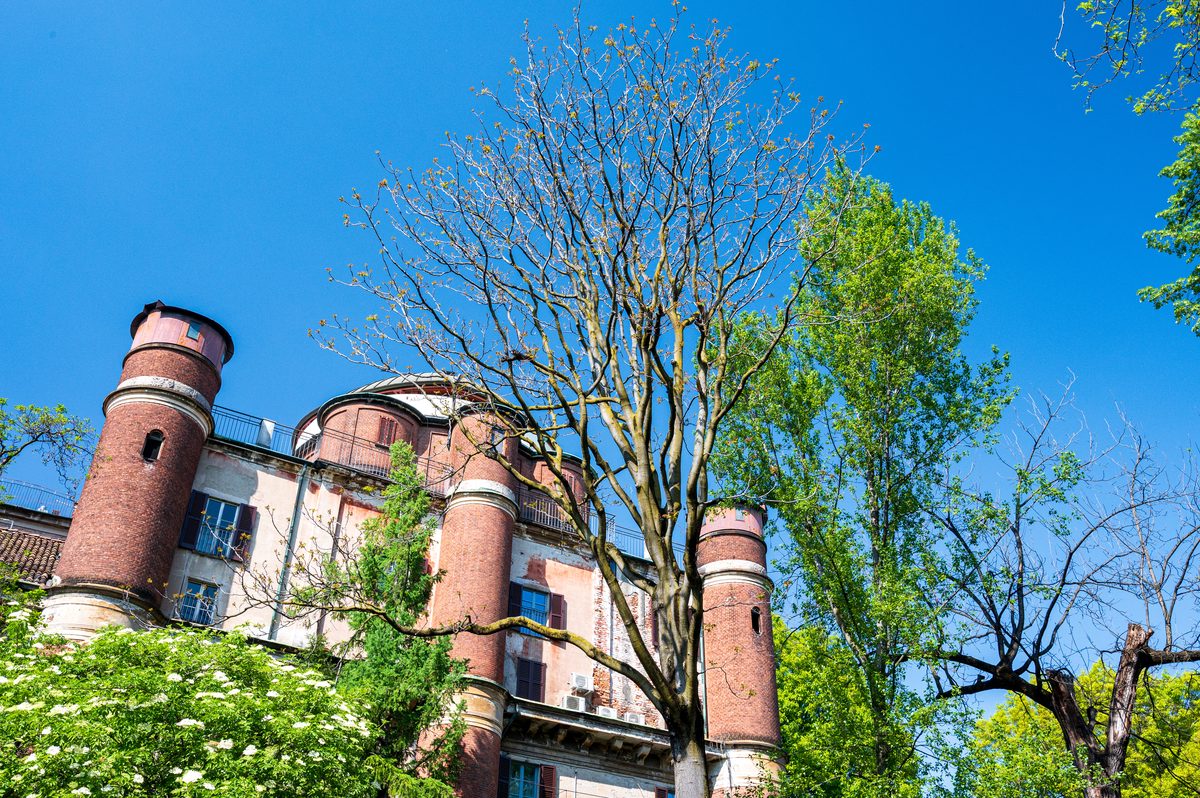 Historic brick towers rise between leafy spring trees beneath an intense blue Milanese sky. The unusual perspective reveals a quieter architectural side of the city, away from its monumental squares and busy streets.