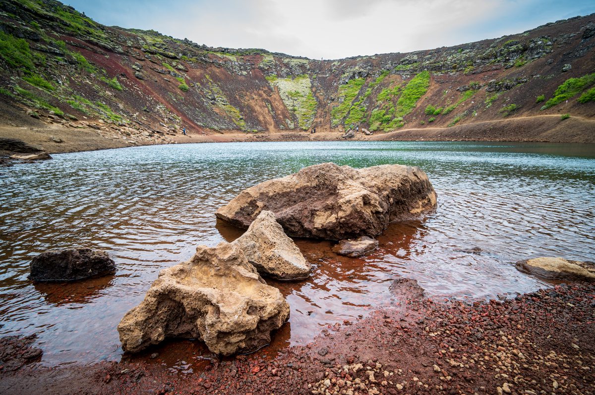 From the water's edge inside a volcanic caldera, textured rocks rest in the shallows of a brilliant aquamarine lake, contrasting sharply with the steep, mineral-rich red walls.
