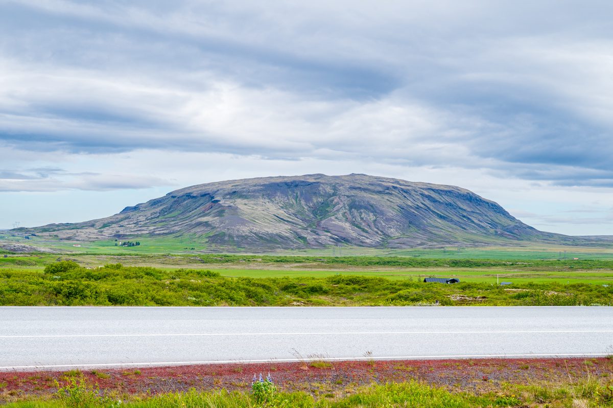 A majestic, sprawling mountain scarred by ancient geological forces anchors the landscape, sitting quietly beyond a vibrant green valley and a deserted stretch of road.