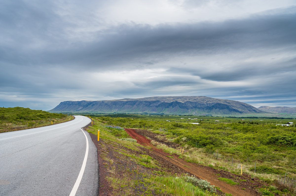 An empty, beautifully paved highway curves gracefully through vibrant green brush, leading the eye directly toward a massive, flat-topped mountain dominating the distant horizon.