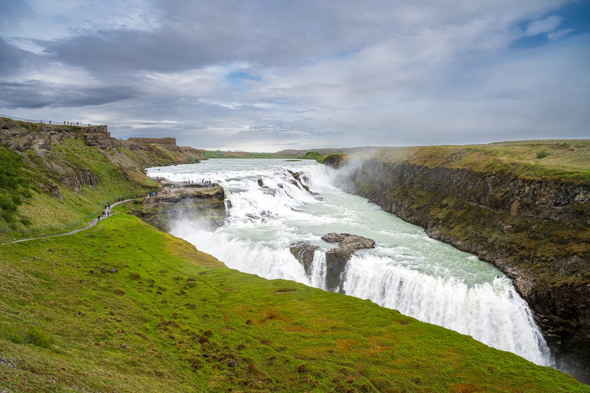A wide perspective of a magnificent, tiered waterfall cascading dramatically through a jagged rocky canyon lined with green slopes and awestruck visitors.