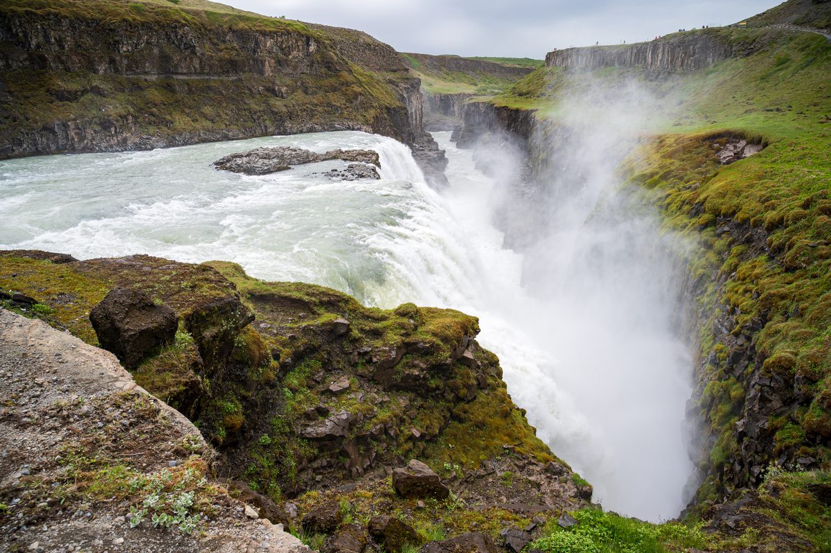 The raw power of nature is on full display as churning glacial waters plummet into a narrow, moss-lined crevice, creating a perpetual wall of mist against the dark rock.