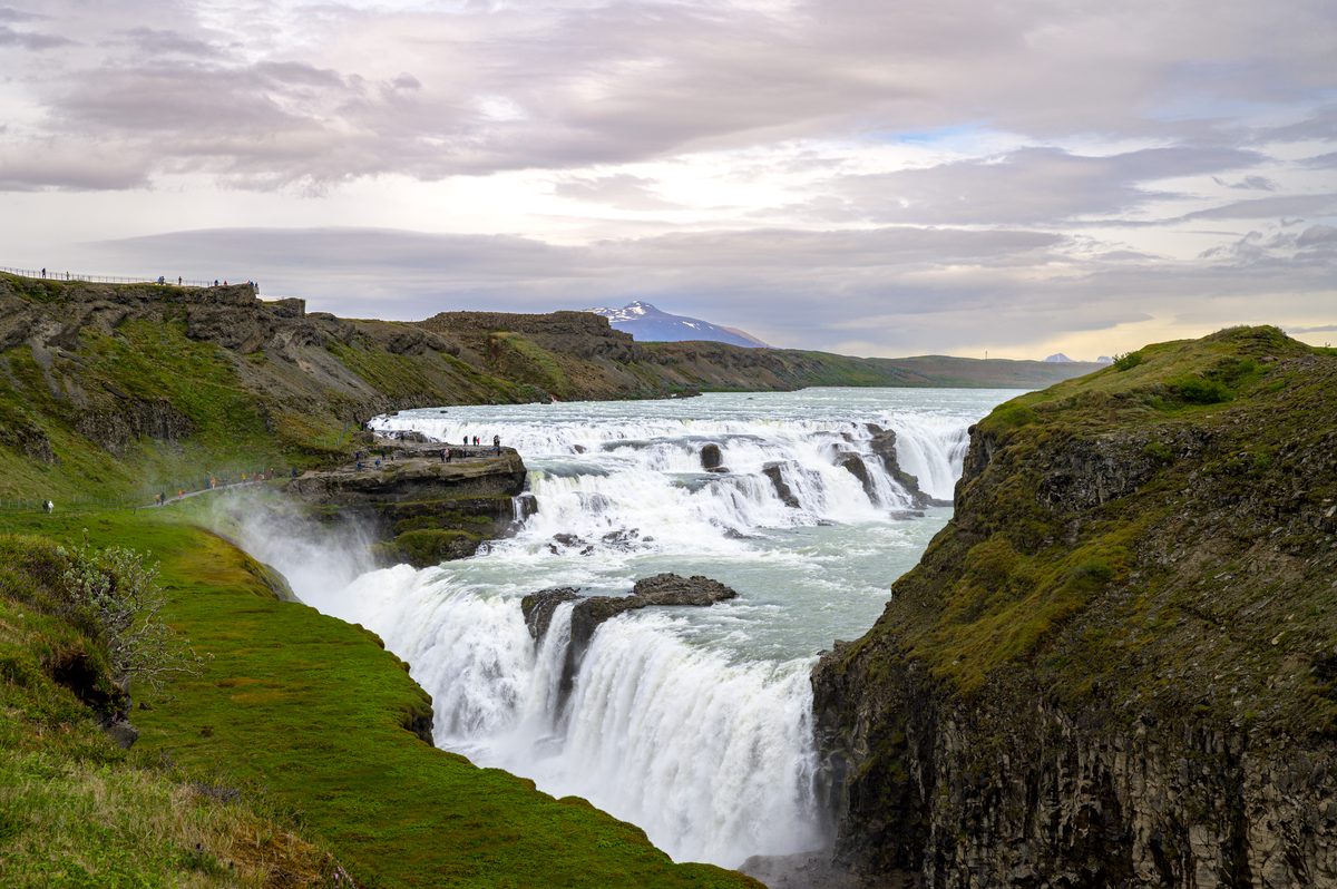 The expansive, tiered cascades of the waterfall flow powerfully through the canyon, framed perfectly by a distant snow-capped mountain peak under a dramatic, cloudy sky.
