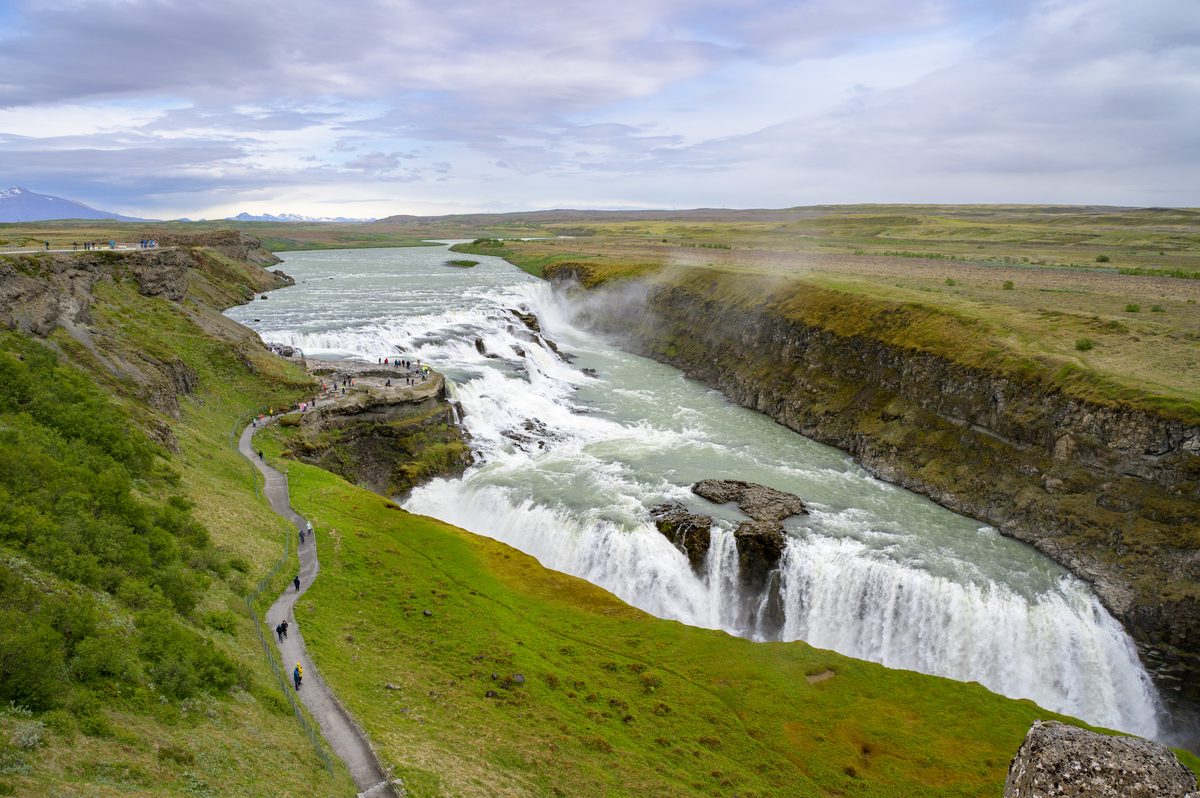 A winding trail leads visitors down a vibrant green hillside to the edge of a roaring river just before it drops violently into a deep, mist-filled gorge.
