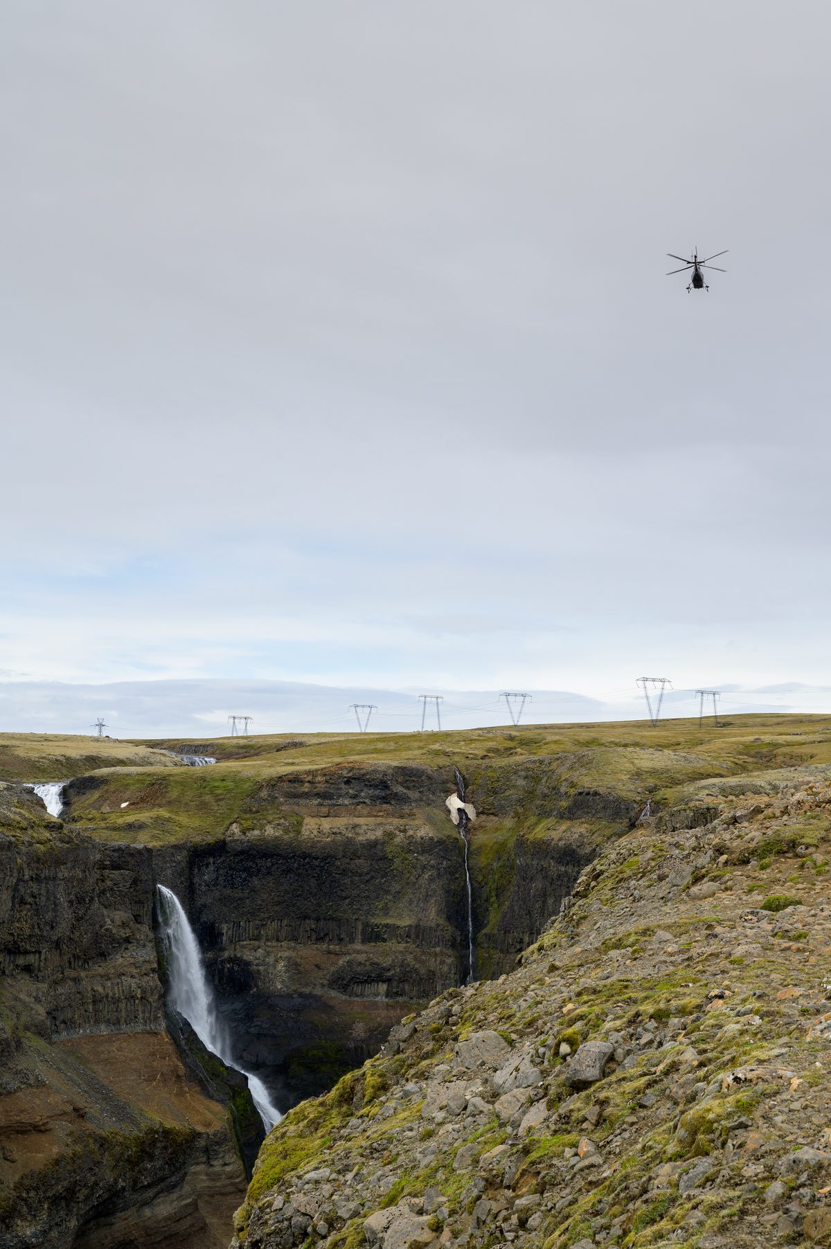 A helicopter hovers high above the deep Haifoss canyon and its cascading waterfalls, while distant power lines trace a path across the desolate, high-altitude plateau.