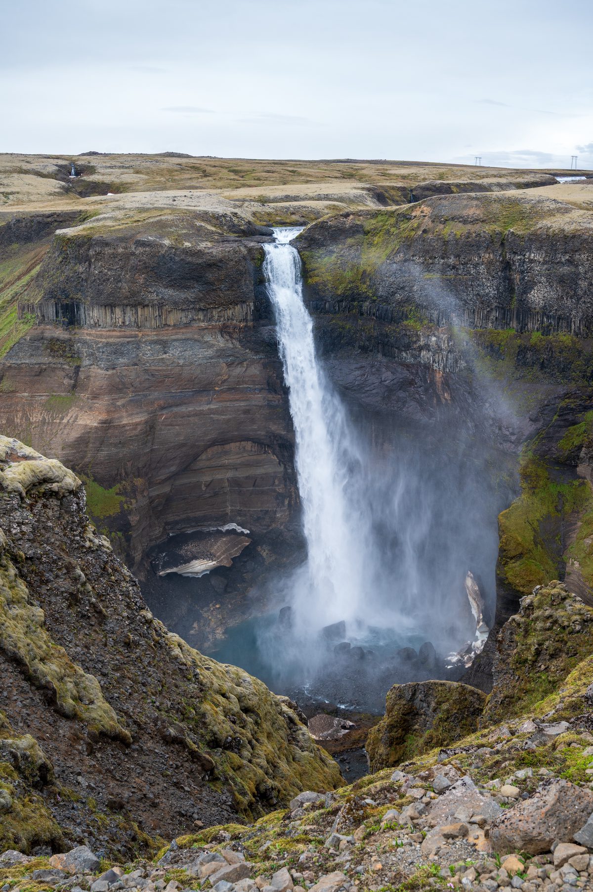 A spectacular, sheer waterfall drops dramatically into a massive, echoing gorge, its thundering waters surrounded by steep cliffs that showcase millennia of volcanic rock layers.