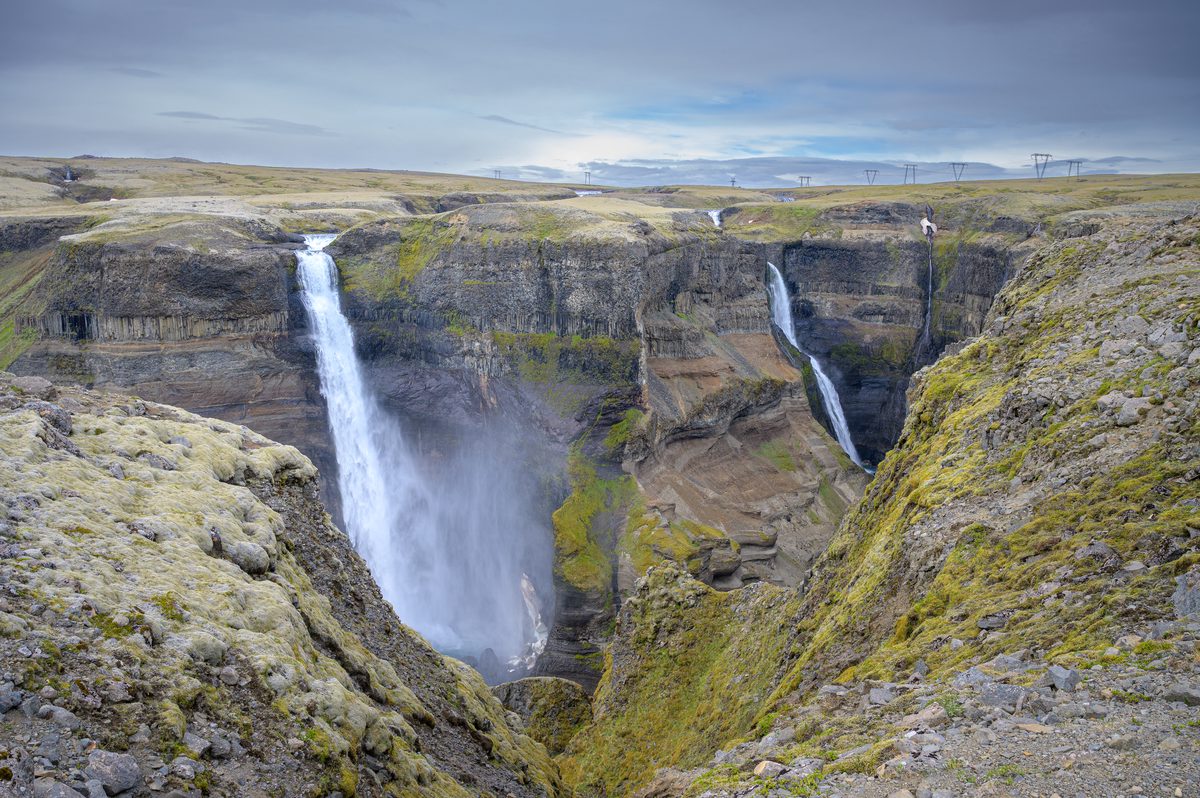 A breathtaking wide view of a massive canyon where two towering waterfalls plunge side-by-side into the misty depths, carving their way through the ancient, moss-draped rock.