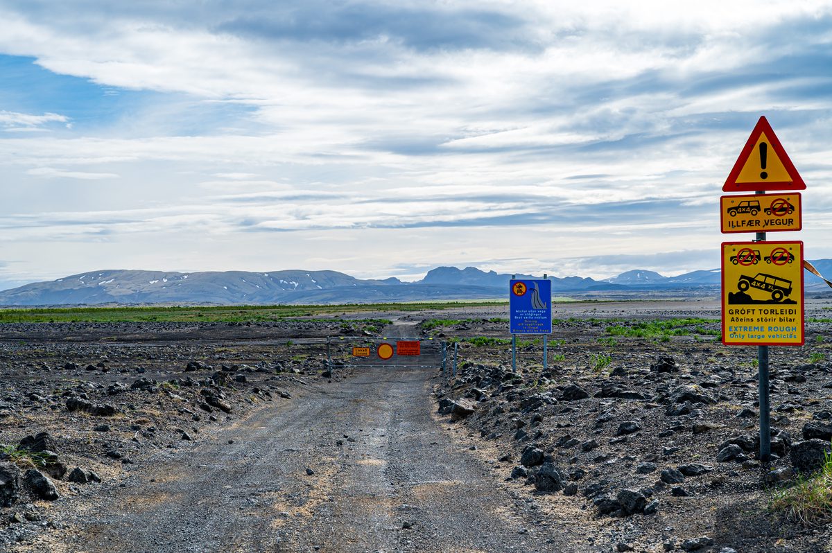 Warning signs mark the entrance to an extreme highland track across the dark lava fields, with distant mountains under a brightening sky.