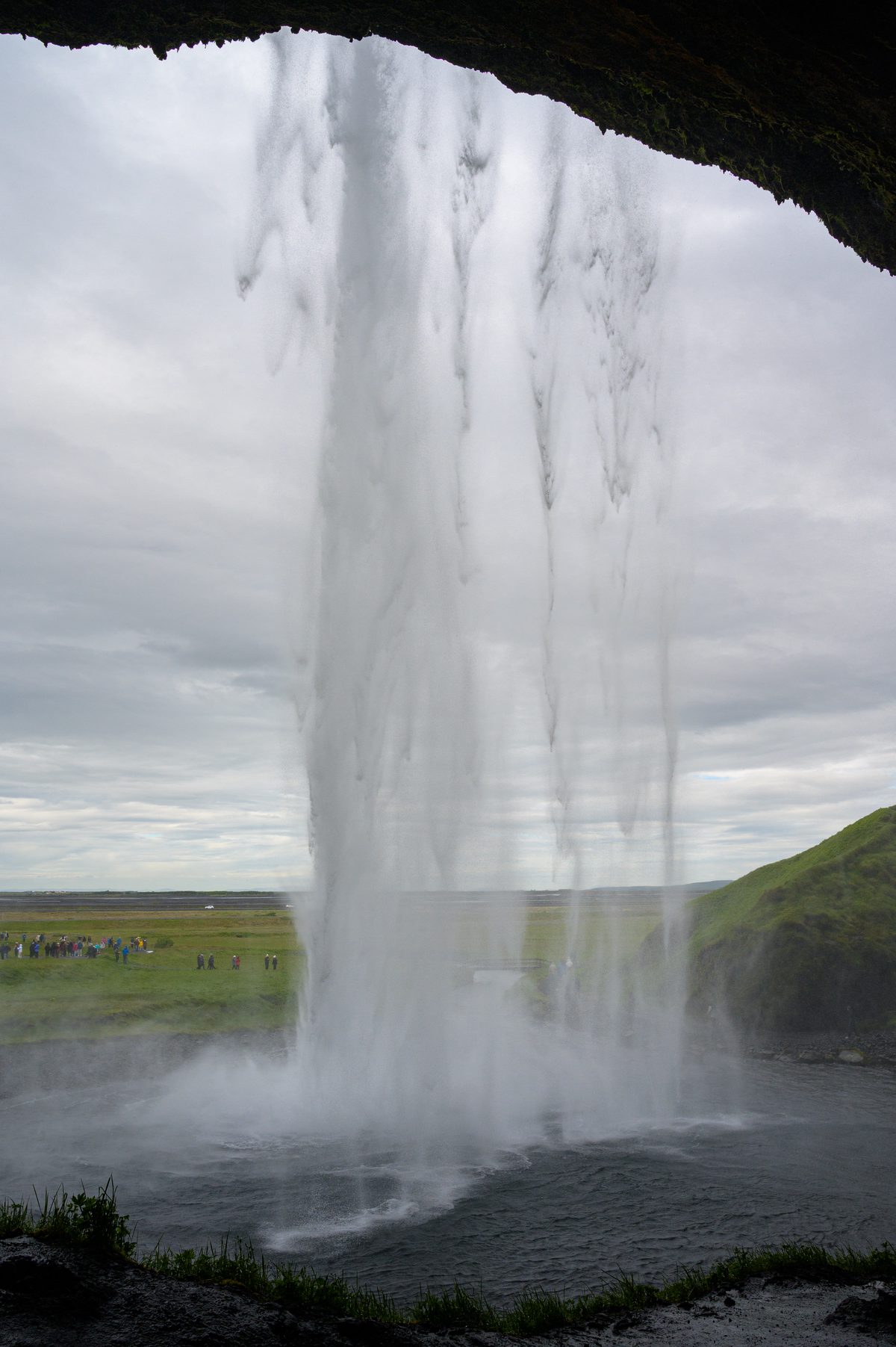 A close view from behind Seljalandsfoss frames the falling water against the distant fields and pale overcast sky.