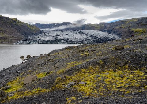 The ash-striped tongue of Sólheimajökull meets a quiet lagoon bordered by black gravel and bright patches of moss.