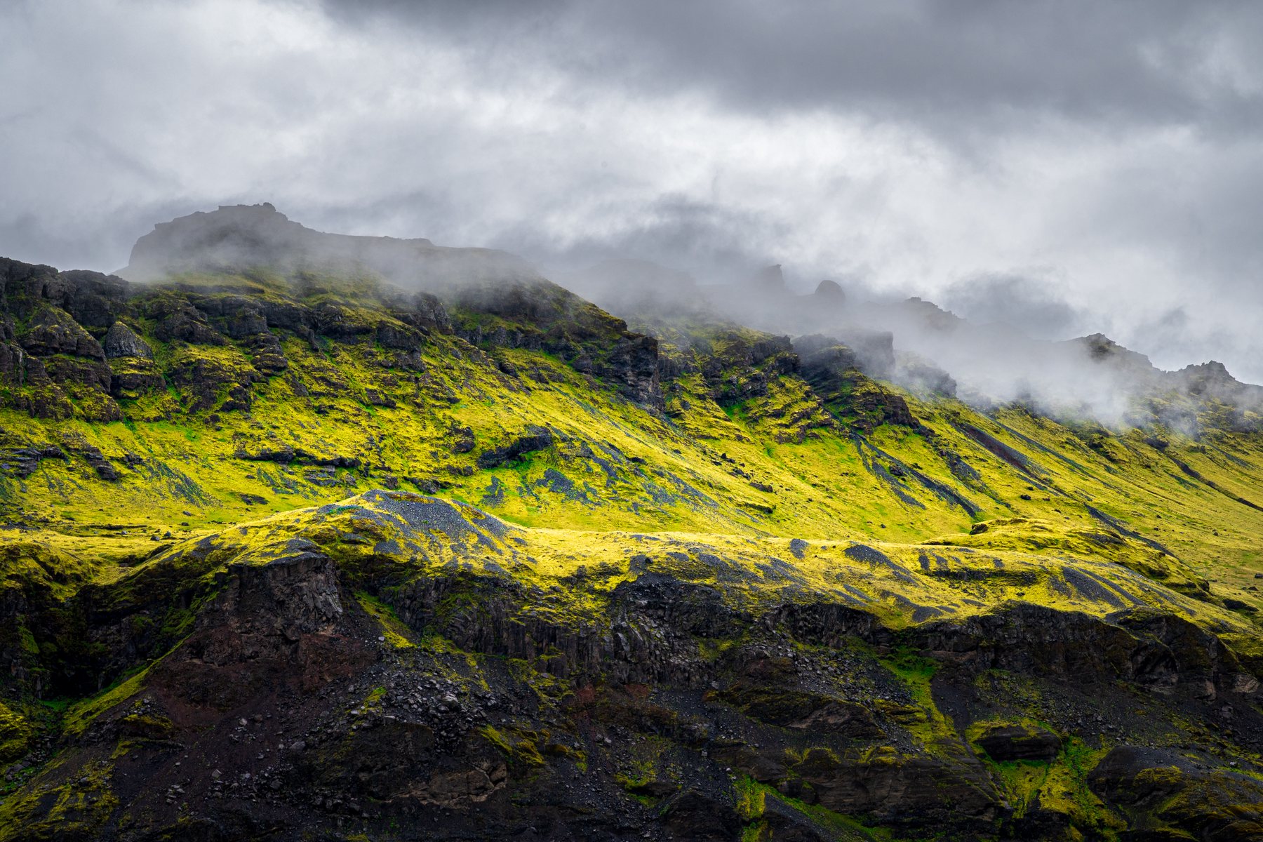 A shaft of light ignites the green slopes above Sólheimajökull while mist curls beneath a dark, restless sky.