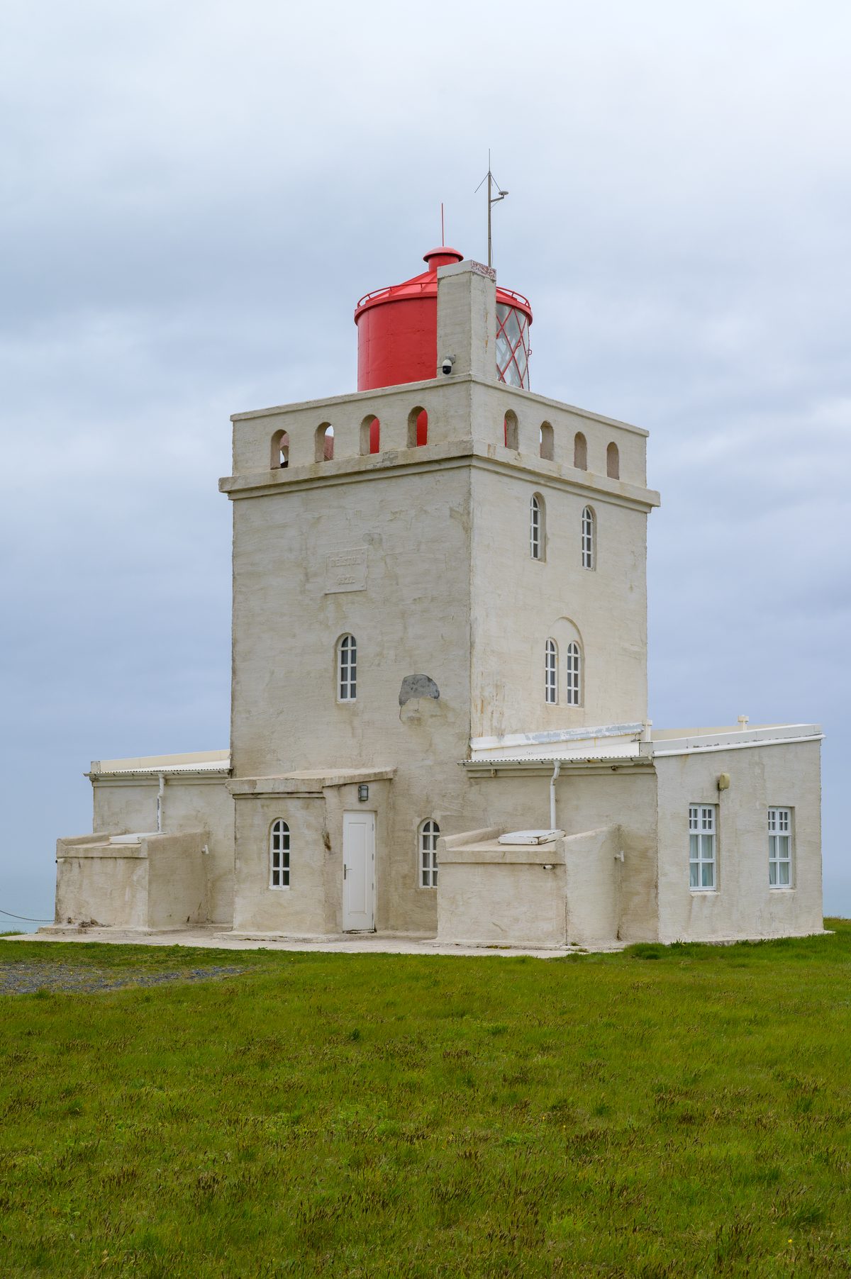 The square white Dyrhólaey lighthouse and its red lantern stand alone on a green headland beneath a soft gray sky.