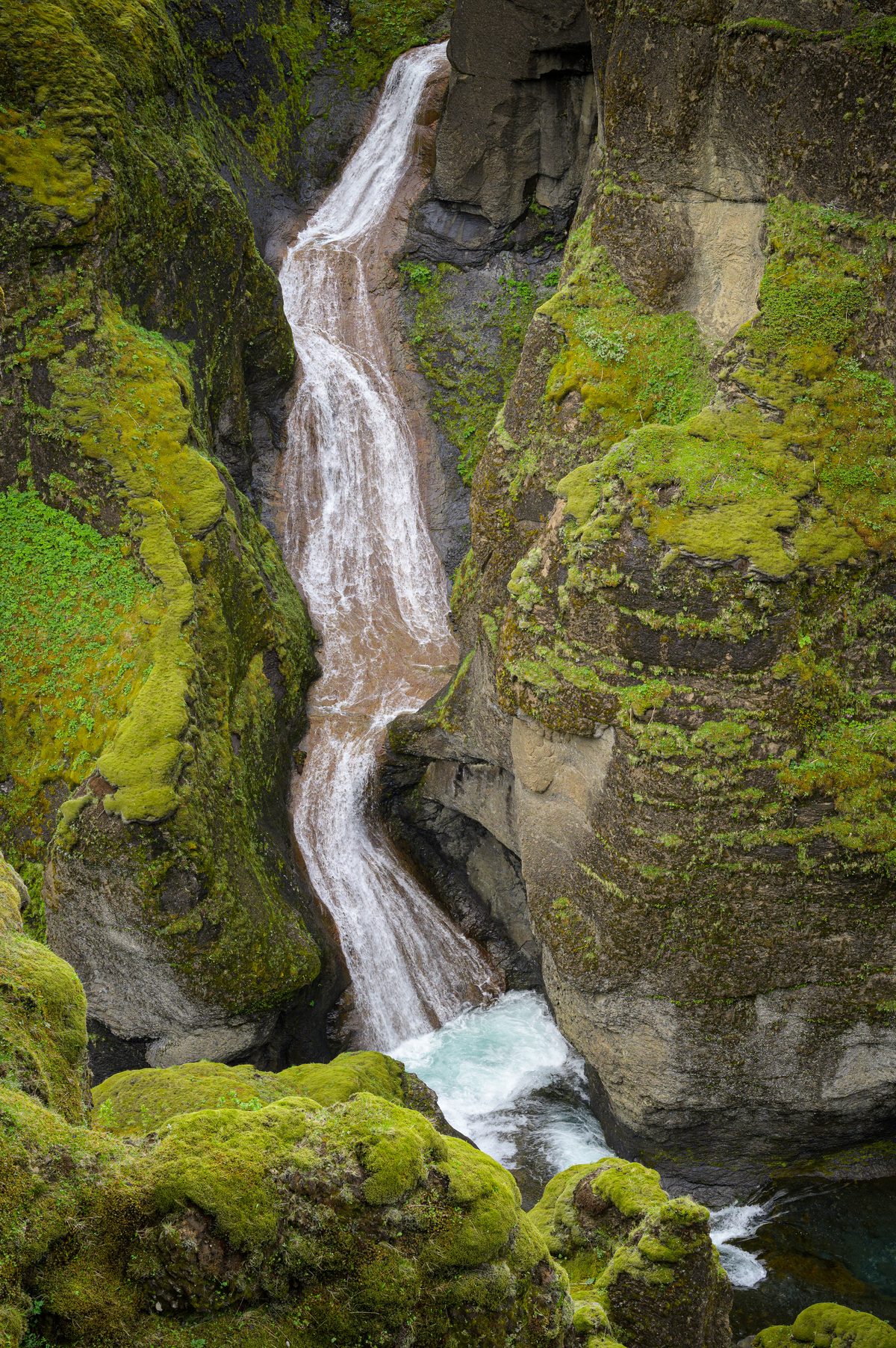 A close view follows rust-tinted water as it tumbles between velvety moss and sheer rock into the river below.