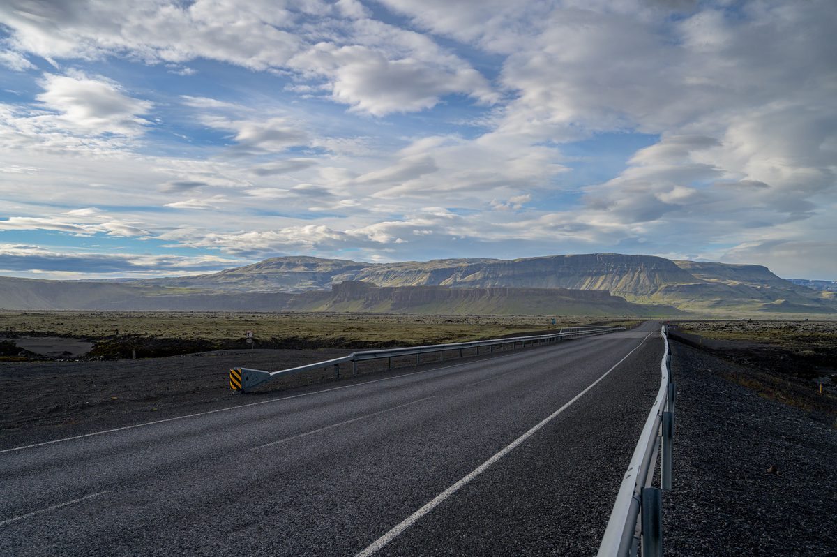 Iceland’s Ring Road runs straight across a bridge toward broad layered mountains beneath a sky breaking into blue.