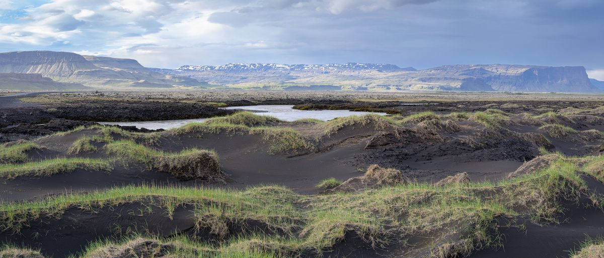 A panoramic view sweeps over grass-tufted black sand, winding water channels and a long wall of mountains lit by passing sun.