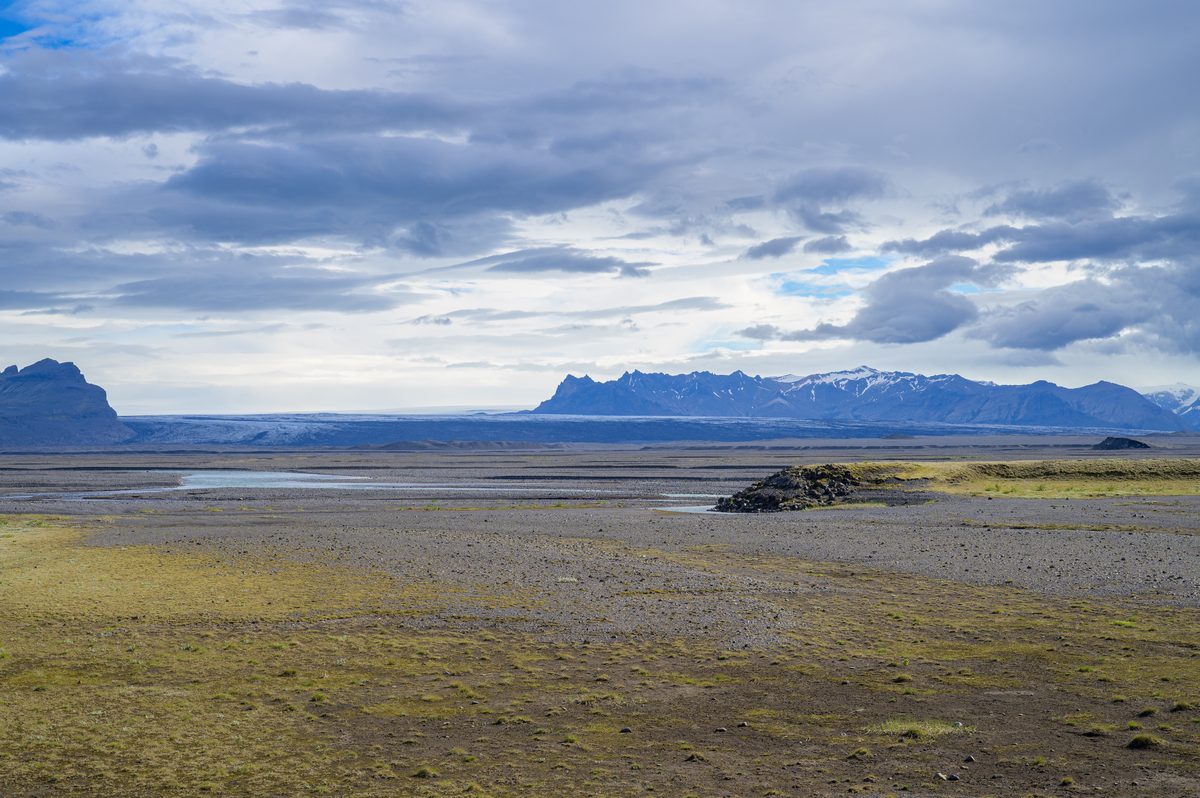 A vast dark outwash plain stretches toward distant jagged peaks beneath a ceiling of silver cloud.