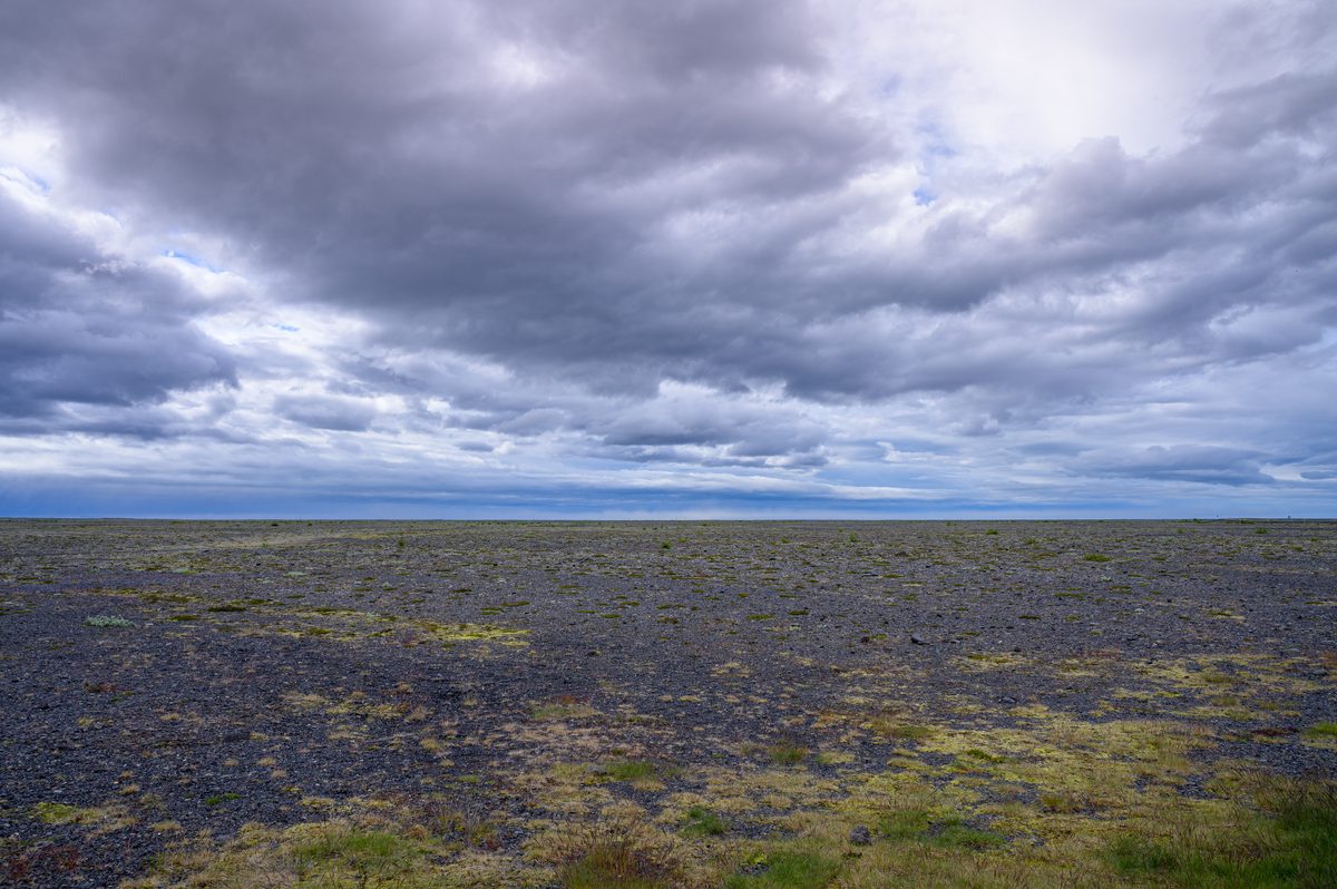 Heavy clouds gather over an immense black glacial plain, broken only by scattered patches of moss.