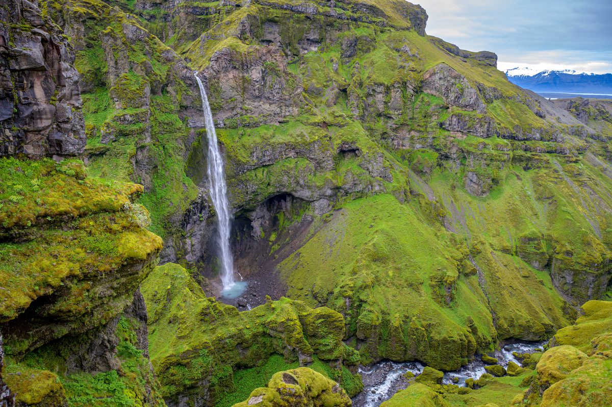 A tall waterfall drops through a vast amphitheater of moss-draped cliffs above a winding stream.