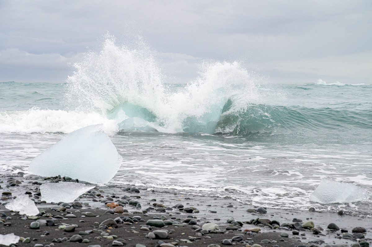 A turquoise wave explodes among stranded ice blocks on the black-pebble shore of Diamond Beach.