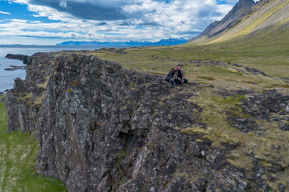 Two travelers pause on the rim of a basalt cliff, with the coast, layered mountains, and distant snowfields stretching behind them.