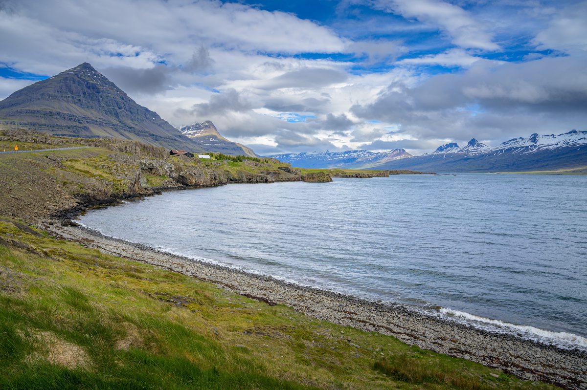 A curved pebble beach meets calm fjord water below pyramidal peaks and a sky breaking into blue.
