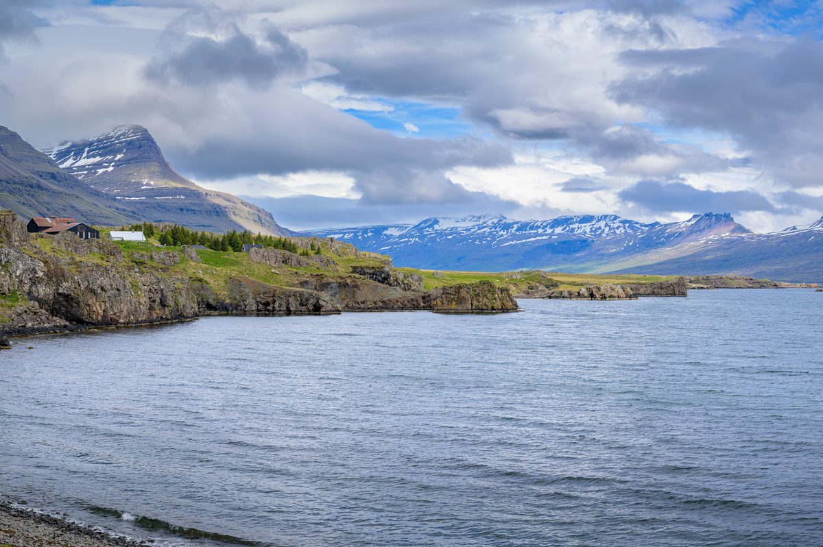 A panoramic view follows the rocky shore toward serrated, snow-streaked mountains under bands of cloud and light.