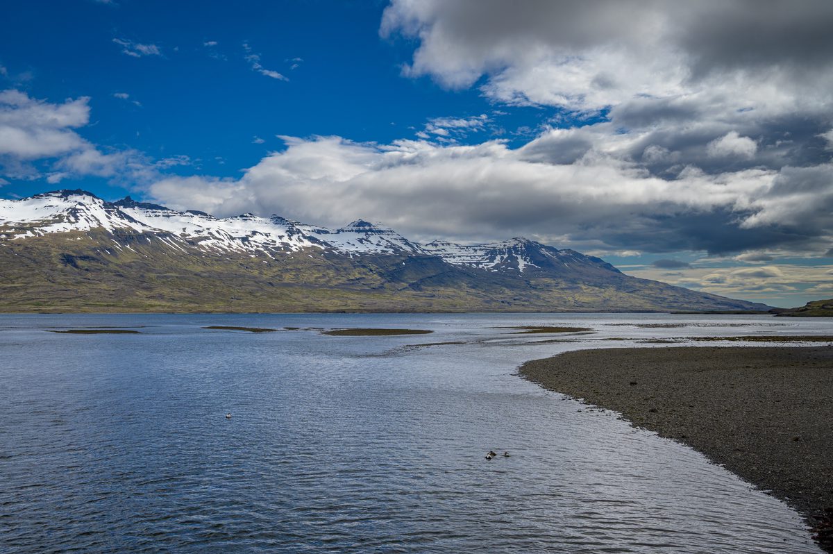 Shallow channels and gravel bars spread across the head of the fjord beneath a long range of snow-covered mountains.