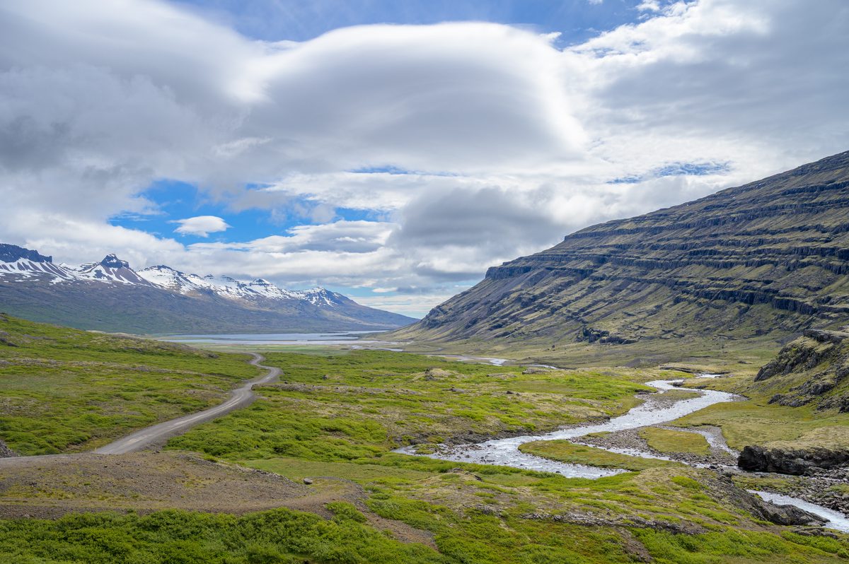 A gravel road and winding stream trace parallel curves toward the fjord beneath sculpted cloud.