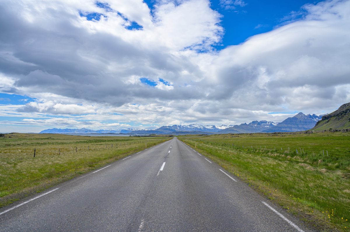 An empty road runs straight across open coastal grassland toward a distant chain of Eastfjord peaks.