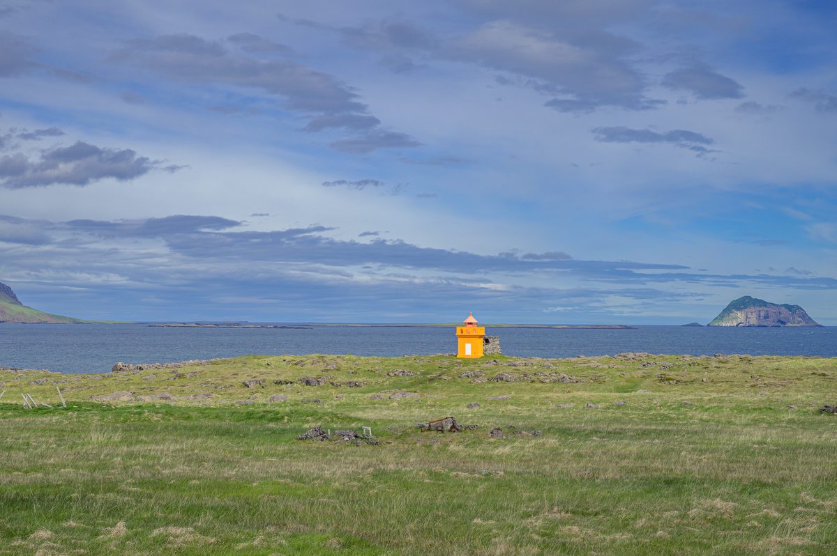 A small orange lighthouse glows against muted grass, blue water, and an isolated flat-topped sea stack.