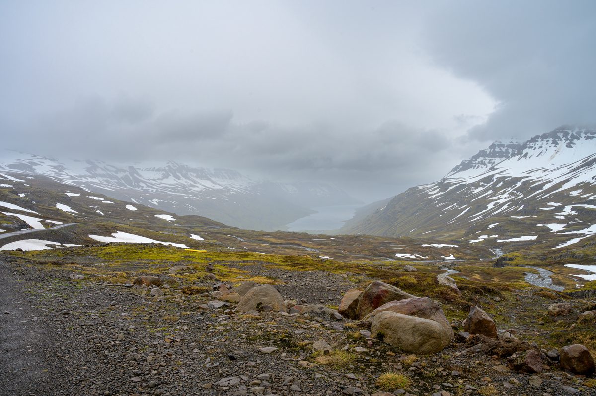 A rough highland track crosses snow-patched ground toward a narrow fjord almost hidden by rain and cloud.
