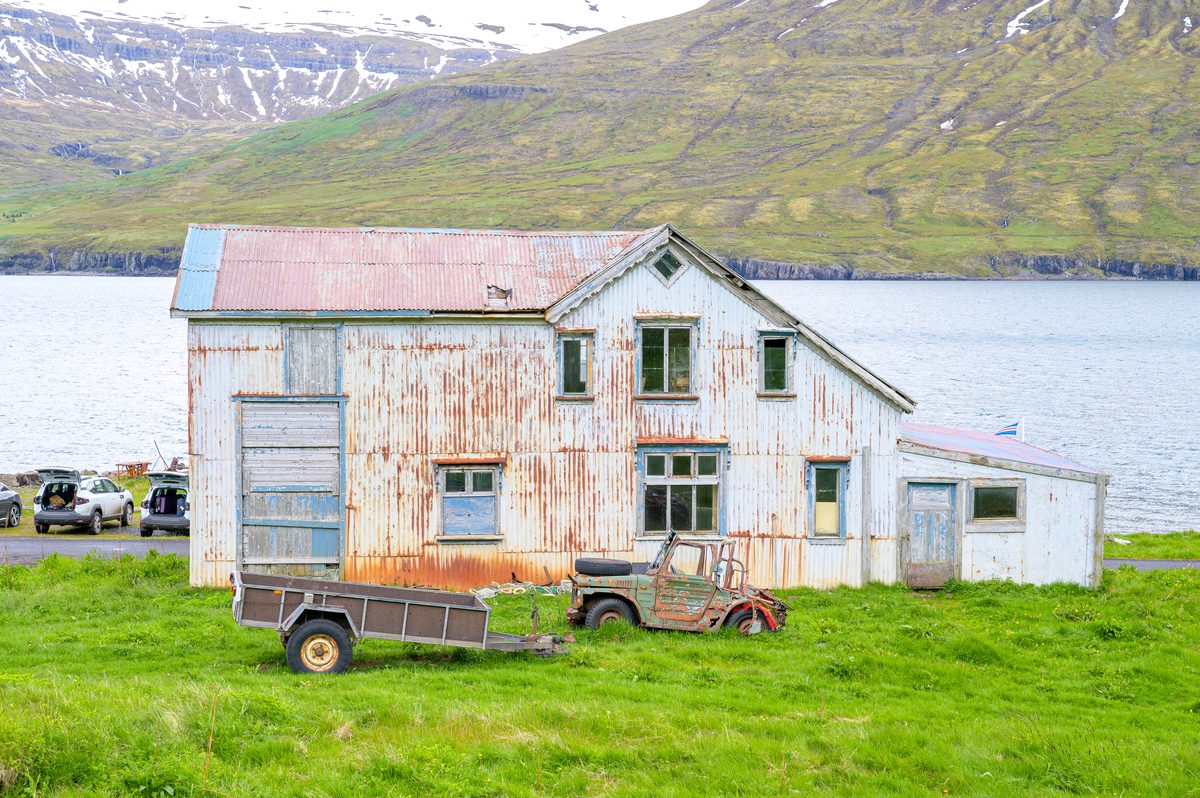 A rust-streaked corrugated workshop and old machinery stand against the quiet water and green mountain slopes.