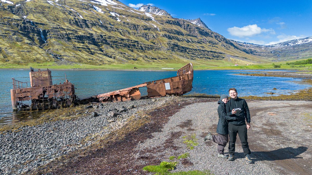 A traveler stands on the stony shore beside the rusted landing craft in Mjóifjörður, with blue water and layered green mountains behind.