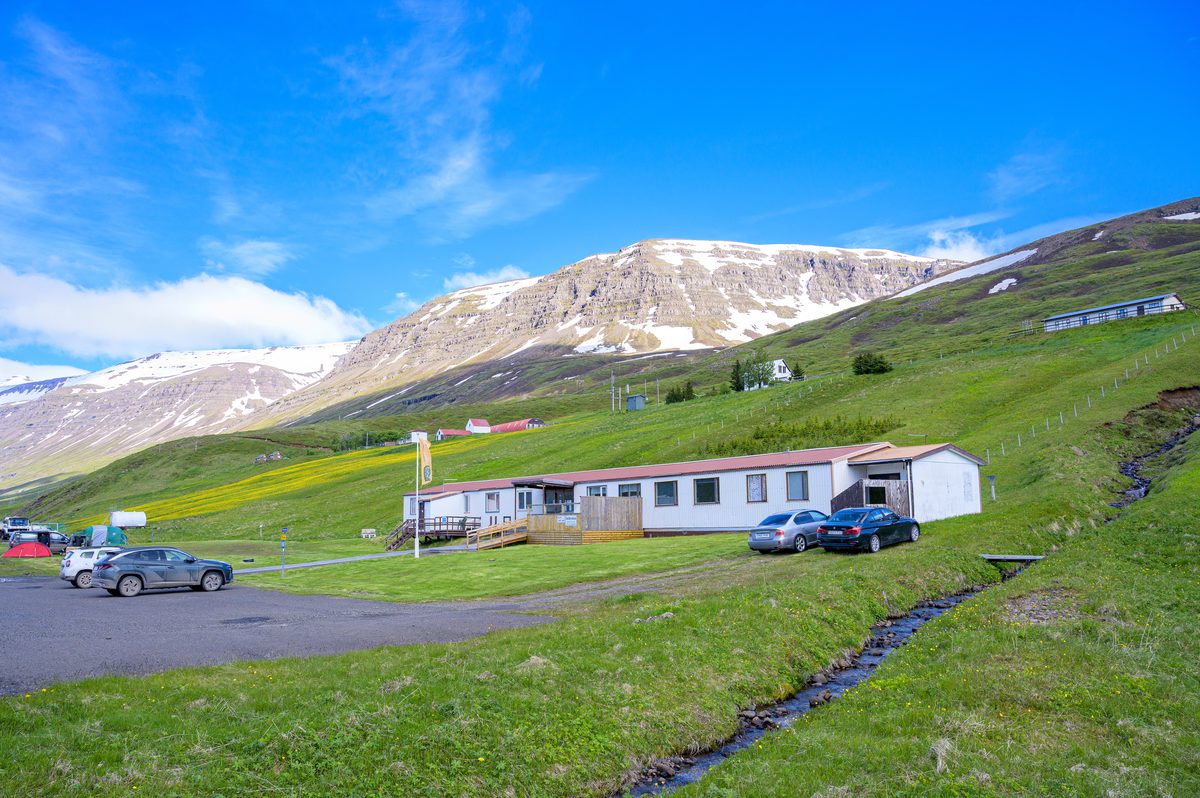 Low houses and parked vehicles sit on a vivid green slope below the pale, snow-streaked mountains of Mjóifjörður.