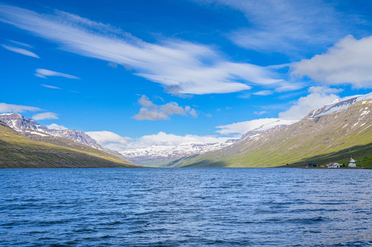 Calm blue water leads into a long mountain valley, with green lower slopes and snowfields glowing in clear summer light.