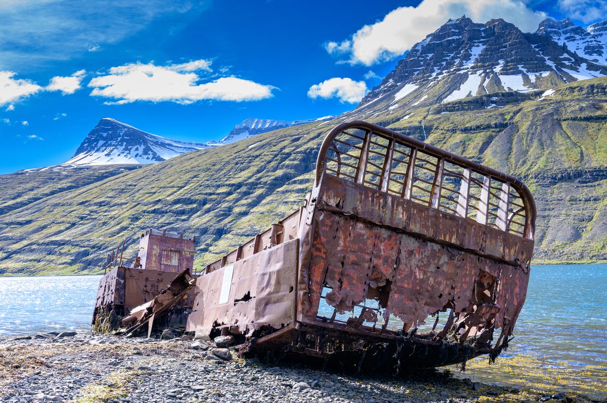 The corroded bow and collapsed hull of an old landing craft rest on black stones beside the blue fjord.