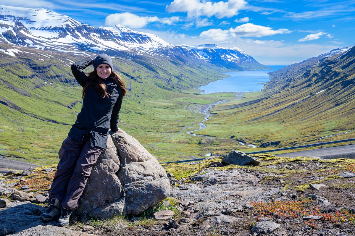 A smiling traveler leans against a roadside boulder with the green valley and distant water of Mjóifjörður below.