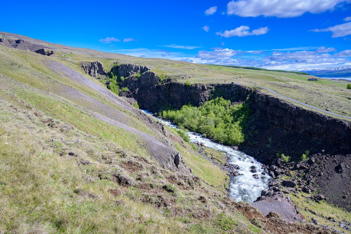 A white stream rushes through a narrow dark ravine carved into a grassy plateau beneath a vivid blue sky.
