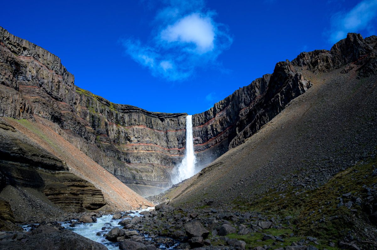 A waterfall pours from a high basalt rim into a shadowed bowl of red scree, black rock, and sparse green grass.