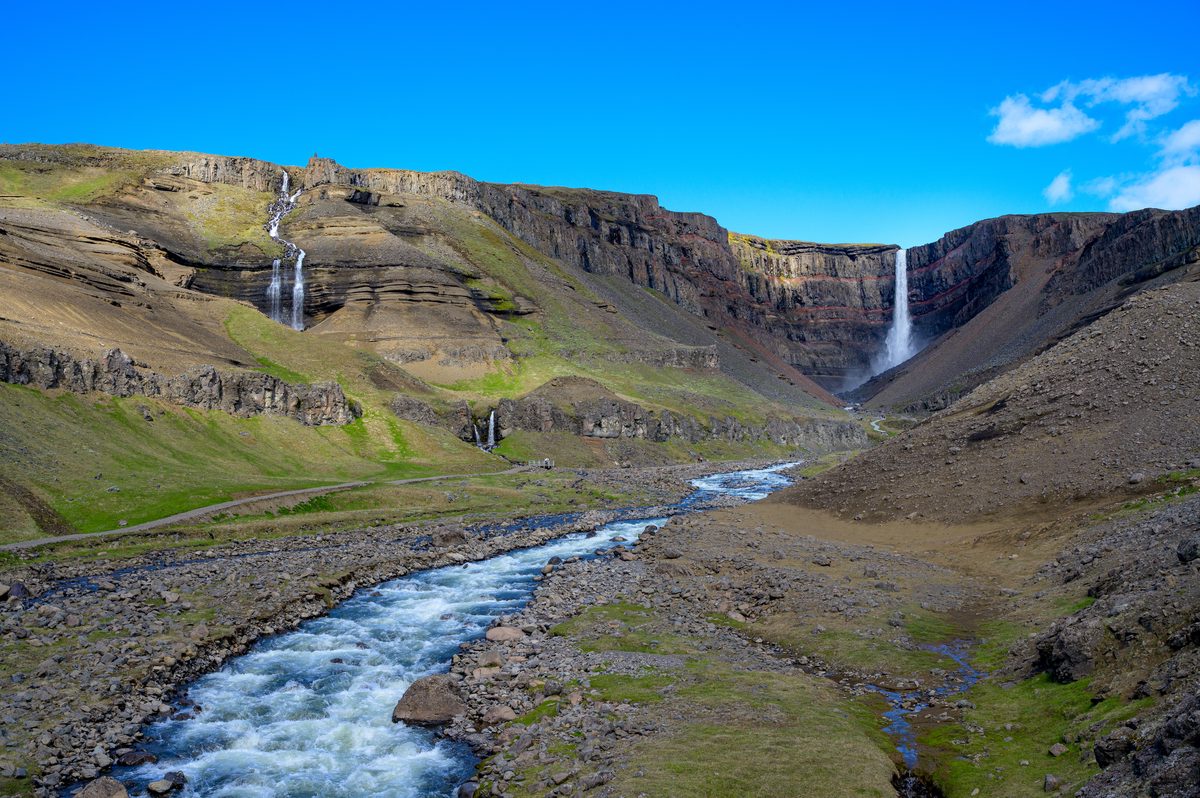 A wide valley view follows a pale river upstream toward a waterfall spilling over a dark cliff band.